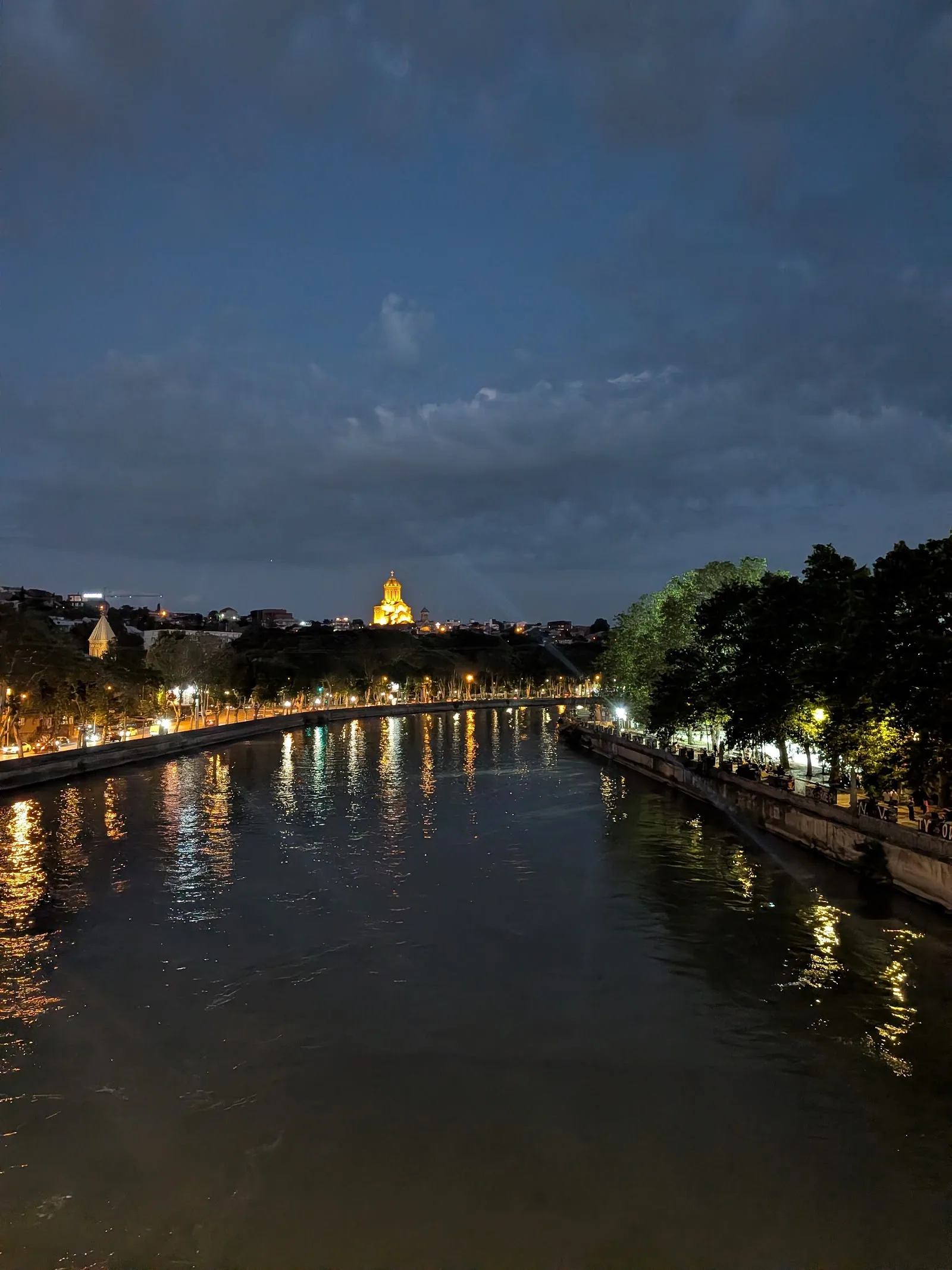 The Mtkvari river at night in Tbilisi with Sameba cathedral lit gold on the hill above.